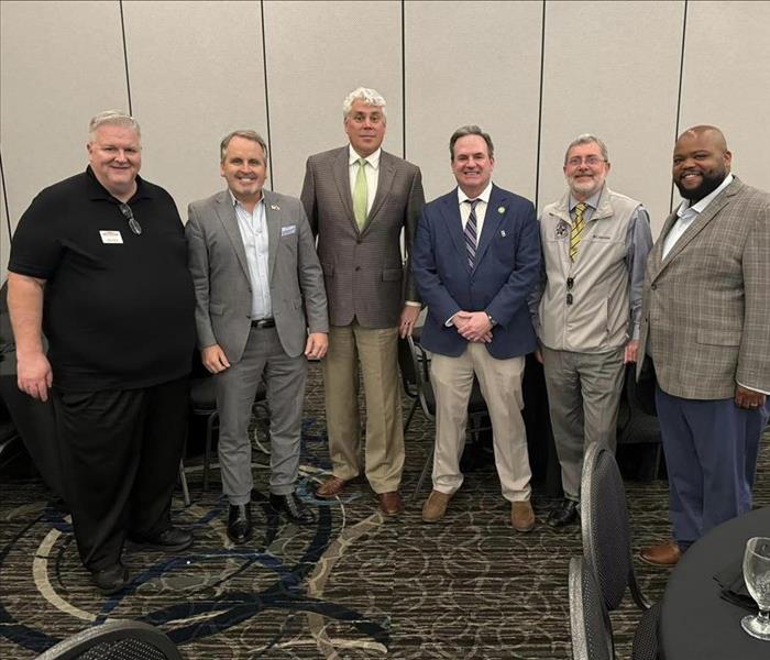 Several men standing in a row smiling for a picture at a luncheon event