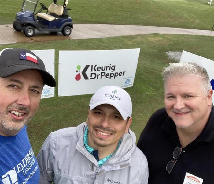 Three men smiling in front of signage for a golf tournament